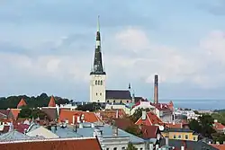 Farbfotografie in der Obersicht von einer hellen Kirche, die von alten Häusern umgeben ist. Die dunkle Turmspitze ist teilweise oxidiert. Im rechten Hintergrund ist das Meer zu sehen.