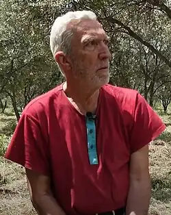 Tayfun Çorağan mit kurzen, weißen Haaren, die ein rotes Hemd trägt. Das Hemd hat eine blaue, durchgehende Knopfleiste. Er befindet sich draußen, inmitten einer Baumgruppe, möglicherweise Olivenbäume und schaut nachdenklich. Sonnenlicht strahlt durch die Blätter und sorgt für einen natürlichen Glanz. Der Boden ist mit trockenem Gras oder Stroh bedeckt, was auf eine ländliche Umgebung hinweist. An dem Hemd ist ein Mikrofon befestigt, was darauf hindeutet, dass die Person möglicherweise ein Interview gibt oder vor einer Kamera spricht. Die gesamte Szenerie wirkt ruhig und idyllisch und vermittelt eine Verbindung zur Natur.