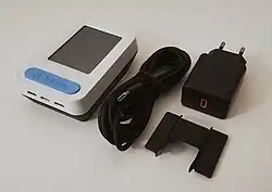 Ein rechteckiges Gerät mit weißem Kunststoffgehäuse und Frontbildschirm samt eines blauen Tastschalters, daneben ein USB-C-Kabel samt Netzteil und Fensteraufhängung