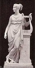 Statue von Terpsichore, Frontalansicht; sie lehnt sich an ein Podest links von ihr, auf dem eine große Lyra steht, diese hält sie zugleich mit der linken Hand