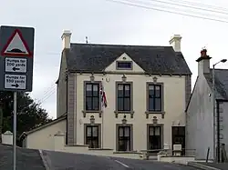 Ein zweistöckiges Gebäude mit hohen Fenstern, einer Plakette im Dachfirst und einer gehissten britischen Flagge davor.