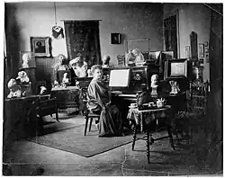 Eine Frau mit einem langen Gewand oder Malkittel bekleidet sitzt an einem Klavier. Ihr Zimmer ist mit Bildern, Wand- und Bodenteppichen eingerichtet. Auf Tischen und Kommoden stehen Büsten und Tonmodelle.