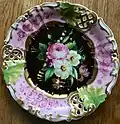Marke: TPM u. preußischer Adler (1847). Kuchenteller, neue Form m. durchbrochenem Rand, Blumenmalerei auf schwarzem Fond, Fahne dreipassig mit rosafarbenen Blättern u. Ranken.