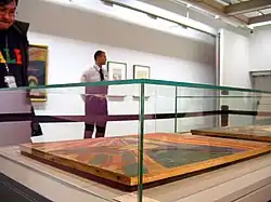 Farbfotografie von einer Seitenaufnahme einer Ausstellungsvitrine, in der das Werk „Der Schrei“ aus dem Jahre 1910 liegend ausgestellt ist. Im Hintergrund stehen ein Besucher und ein Museumswärter. An der hinteren Wand sind weitere Gemälde zu sehen.