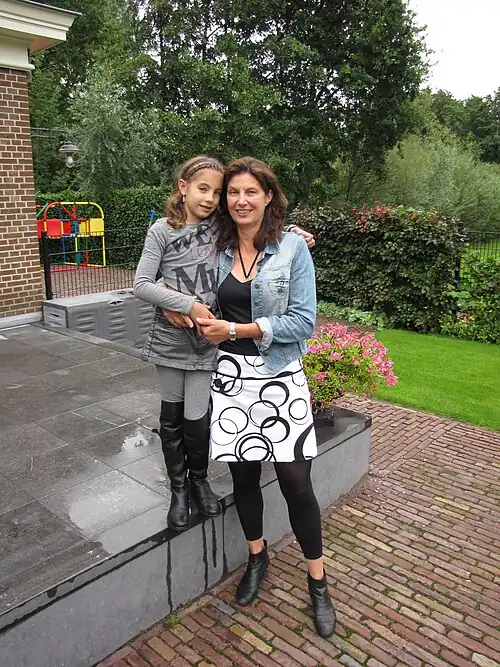 Tineke Hidding und ihre Tochter stehen im Garten ihres Hauses. Tineke Hidding trägt eine blaue Jeansjacke über einer schwarzen Bluse zu einem weißen Rock, die Tochter trägt graue Kleidung.