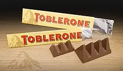 Zwei offene Toblerone-Packungen mit einem groß- und einem kleinzackigen Tobleroneriegel. Zu sehen ist noch das alte Toblerone-Logo in Form des Matterhorns mit einer Schattensilhouette des Berner Bärs auf den Packungen und dem Tisch darunter.