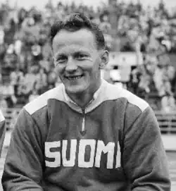 Schwarzweißfoto: Toivo Hyytiäinen sitzt im Trainingsanzug mit der weißen Aufschrift Suomi.