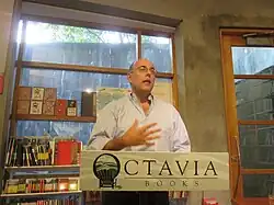 Autor stehend, sichtbar ab Brustkorb vor einem Fenster mit einem Bücherregal in Farbe.