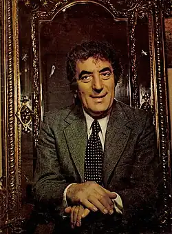 Männliche Person im mittleren Alter von vorn sitzend im grauen Anzug, weißem Hemd und dunkler weiß gepunkteter Krawatte. Er scheint auf einem kunstvoll gestalteten thronartigem Stuhl mit weinroter Rückenlehne eingerahmt von goldenen Ornamenten zu sitzen.