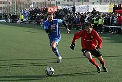 Frontale Farbfotografie von zwei Fußballspielern, die einem Ball in der Bildmitte hinterherjagen. Der linke Spieler trägt ein blaues Trikot und der rechte Spieler ein rotes Trikot und schwarze Torwarthandschuhe. Im Hintergrund steht ein kleines Publikum.