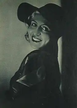 Seitlich stehende Traute Flamme lächelt offen und trägt einen großen, breiten Hut. Der Hut ist dunkel und scheint aus einem festen Material zu bestehen. Sie trägt zudem einen langen, glänzenden Mantel, der ebenfalls dunkel ist. Der Mantel reflektiert das Licht leicht, was auf ein glattes oder glänzendes Material hinweist. Der Hintergrund des Bildes ist dunkel und leer, ohne erkennbare Details oder weitere Objekte. Das Schwarz-Weiß-Bild vermittelt insgesamt eine geheimnisvolle und formelle Atmosphäre.