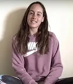 Mas sitzt vor einer weißen Wand. Sie trägt eine blaue Trainingshose und einen lila Pullover.