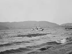 Schwarz-Weiß-Foto eines kleinen Schiffes. Am Bildrand ist ein flaches Ufer und ein Bootssteg erkennbar.
