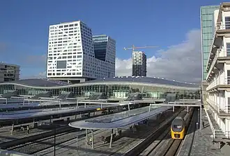 Moderner Bahnhof Utrecht Centraal mit geschwungenem Glasdach und überdachten Bahnsteigen, ein gelb-schwarzer Zug von Nederlandse Spoorwegen fährt rechts ein. Im Hintergrund Hochhäuser, darunter ein weißer Turm mit diagonalen Linien, sowie ein Baukran vor blauem Himmel.