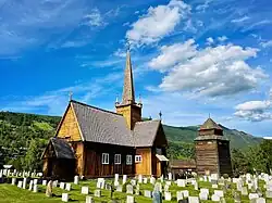 Foto einer Holzkirche mit Grabsteinen im Vordergrund und einer Erhebung dahinter