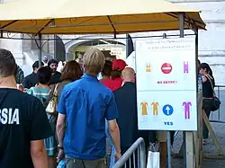 Touristen am Petersdom. Die piktographische Hinweistafel verdeutlicht die erwünschte Kleiderordnung. Inakzeptable Kleidungsstücke lassen die Schultern und Knie frei, bei den als akzeptabel geltenden sind Knie und Schultern bedeckt