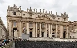 Frontale Farbfotografie von der Fassade eines Renaissance-Gebäudes mit vielen Stuhlreihen und großer Menschenmenge im Vordergrund. Vor dem Gebäude befinden sich die Marmorfigur eines Mannes in griechischem Gewand und ein weißes, überdachtes Podium. Das dreigeschossige Gebäude hat unter dem Giebel eine lateinische Inschrift und mehrere Säulen. Die Dachkante ist mit einigen Figuren bestückt. Links und rechts sind zwei Uhren mit Glocken darunter.