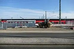 Ein gelbes Baustellenfahrzeug steht vor Absperrgittern. Im Hintergrund sieht man eine rote S-Bahn.