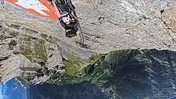 Kletterer am Leukerbad-Klettersteig auf Höhe der ikonischen Metalltafel mit der Schweizer Flagge, aus etwa 2320&nbsp;m Blick herab auf das darunter gelegene Leukerbad