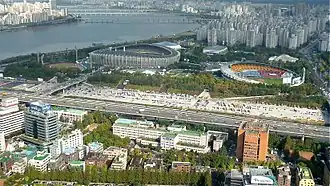 Blick auf das Olympiastadion und weitere Sportstätten im Seoul Sports Complex. Erkennbar ist die Lage nahe am Ufer des Hangang Fluss. Das Gelände liegt inmitten von Schnellstraßen und dichter urbaner Bebauung.
