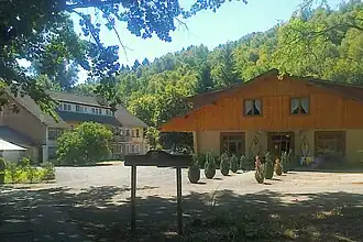 Das Foto zeigt den teilweise durch Bäume beschatteten Hof eines Anwesens bei sonnigen Wetter. Zwei Gebäude sind zu sehen, rechts eines im Chaletstil mit Erdgeschoss und ausgebautem Dachgeschoss, links ein größeres mit zwei Geschossen zuzüglich ausgebautem Dachgeschoss unter einem Satteldach. Hinter den Häusern steigt ein bewaldeter Hang an.