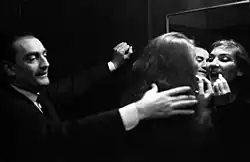 Maria Callas schwarz-weiß Aufnahme trägt vor einem menschhohen Spiegel mit einem Lippenstift Ihre Lippen nach. Luchino Visconti steht link von Ihr mit einer Hand an der Wand gelehnt. Seine rechte Hand liegt über ihrem langen Haar auf ihrer Schulter. Beide Gesichter spiegeln sich in dem Spiegel. Das Foto wurde im Teatro alla Scala in Mailand 1959 aufgenommen.