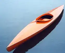 Schlanker, rotbrauner Holzkajak mit offenem Cockpit, liegt unbewegt auf ruhigem Wasser.