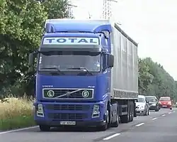 Photo eines fahrenden Sattelzuges, an dessen Sattelzugmaschine vorne eine Plakette mit der Aufschrift „S“ für „supergrünes und sicheres“ Fahrzeug und eine Tafel mit dem Buchstaben&nbsp;L angebracht ist.