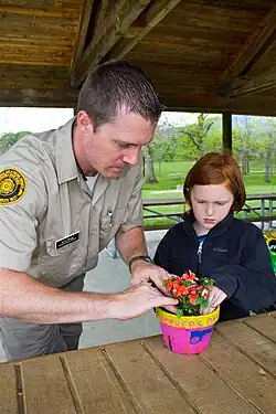 Ein Mann in Uniform und ein rothaariges Kind setzen zusammen eine rot blühende Pflanze in einen Blumentopf, auf dem „Mother's Day“ steht.