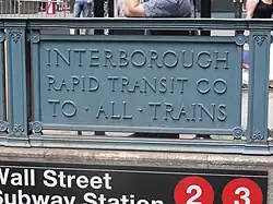 Detail eines Bahnhofseingang: Ein Blechschild als Teil der Brüstung, mit der dreizeiligen Inschrift „Interborough / Rapid Transit Co / To All Trains“