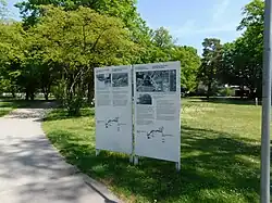 Das Foto zeigt 2 Texttafeln auf dem „Weg des Erinnerns“ in Dachau, die Tafeln stehen in der grünen Wiese, ein Weg führt daran vorbei
