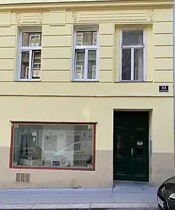 Frontalansicht auf ein Wohnhaus mit drei Fenstern im Obergeschoss und einem Schaufenster sowie dem Hauseingang im Erdgeschoss