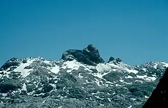 Das Wildalmkirchl mit seiner auffälligen Gestalt in Form einer Kirche. Links unterhalb mit rotem Pfeil markiert ist die Wildalmkirchl-Biwakschachtel (2457 m)