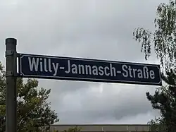 Blaues Straßenschild mit weißer Schrift. Aufschrift: Willy-Jannasch-Straße. Im Hintergrund sind links und rechts Bäume zu sehen. Unten ist das Dach eines Plattenbaus abgebildet.
