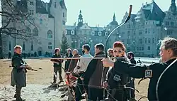Das Foto zeigt aufgereihte Bogenschützen auf einem sonnigen Schlosshof, darunter eine Frau mit bemaltem Gesicht, die direkt in die Kamera blickt.