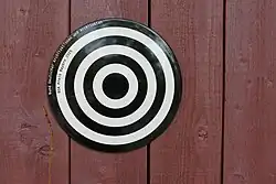 Auf roten Fassadenlatten aus Holz ist ein rundes Metallschild mit schwarz-weißen konzentrischen Kreisen zu sehen. Auf den äußersten beiden Ringen ist die Beschriftung "Bund Deutscher Architektinnen und Architekten. BDA Preis Bayern 2025" angebracht