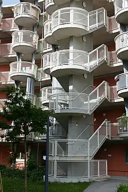 Foto einer Fassade aus rötlichem Holz, das weitgehend von Balkonen und einer Außentreppe aus grauem Metall verdeckt ist