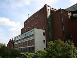 Großes. schlichtes Backsteingebäude mit wenigen Fenstern, vorne angesetzt ein hellerer, schmaler Betonbau mit großen, quadratischen Feldern, die teilweise mit Backstein und Fenstern ausgefüllt sind. Weiter hinten schließt eine ältere Ziegelfassade mit Satteldach an.