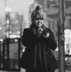 Schwarz-Weiß Fotografie einer stehenden Frau in weitem Mantel, die einen Fotoapparat vor sich in der Hand trägt. Die Frau steht frontal zum Betrachter, nur ihr Oberkörper ist im Bild, ihre Haare sind zu einem lockeren Knoten hochgesteckt.