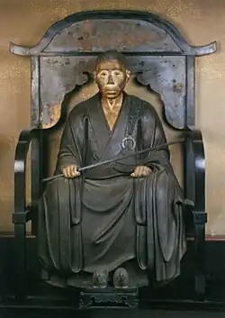 Kunstwerks des sitzenden Mönchs von vorne auf einer Art Thron mit einem Stab in der Hand in Farbe.