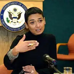 Salbi spricht in ein Mikrophon, sie hat kurze schwarze Haare, trägt einen schwarzen Pullover, sie macht erklärende Gesten mit beiden Händen, im Hintergrund ist das Emblem des US-Außenministeriums sichtbar