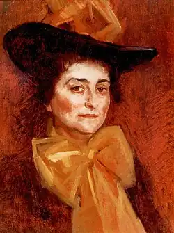 Das Gemälde zeigt vor einem orangen Hintergrund eine Frau mit Hut und einer großen ebenfalls orangen unter dem Kinn gebundenen Schleife, die im Halbprofil zum Betrachter blickt.