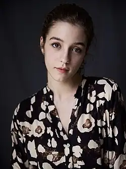 Zoe Bonafonte mit braunen Haaren und einem neutralen Gesichtsausdruck, trägt ein schwarzes Hemd mit auffälligem Muster. Auf dem Hemd befinden sich große, unregelmäßig geformte Flecken in Weiß- und Brauntönen, die an ein abstraktes oder animalisches Design erinnern. Es hat einen V-Ausschnitt mit Knöpfen vorne. Der dunkle Hintergrund hebt die helleren Elemente des Hemdes hervor, wodurch das Muster besonders gut zur Geltung kommt. Insgesamt ist das Hemd ein Blickfang durch sein markantes und einzigartiges Design.