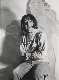 Das schwarz-weiß Foto zeigt Maria Lassnig mit verschränkten Armen in die Kamera schauend. Sie trägt einen hellen Pullover mit einer Aufschrift. Ihr Haar ist halblang und im Hintergrund befindet sich eine Malerei von ihr, die sie mit einer Kamera zeigt.