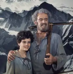 Zarli Carigiet mit Armbrust auf der Schulter in der Rolle des Wilhelm Tell, rechter Art auf der Schulter seines Sohnes, beide in grauem Kapuzenhemd blicken an der Kamera vorbei, Farbfotografie, quadratisch