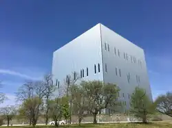 Blick vom Boden auf ein modernes, kubisches, mehrstöckiges Gebäude. Es erscheint silbrig glänzend mit senkrechten, langen Aluminiumpaneelen verkleidet und hat wenige, schlitzartige Fenster. Es steht in einer Grünanlage.
